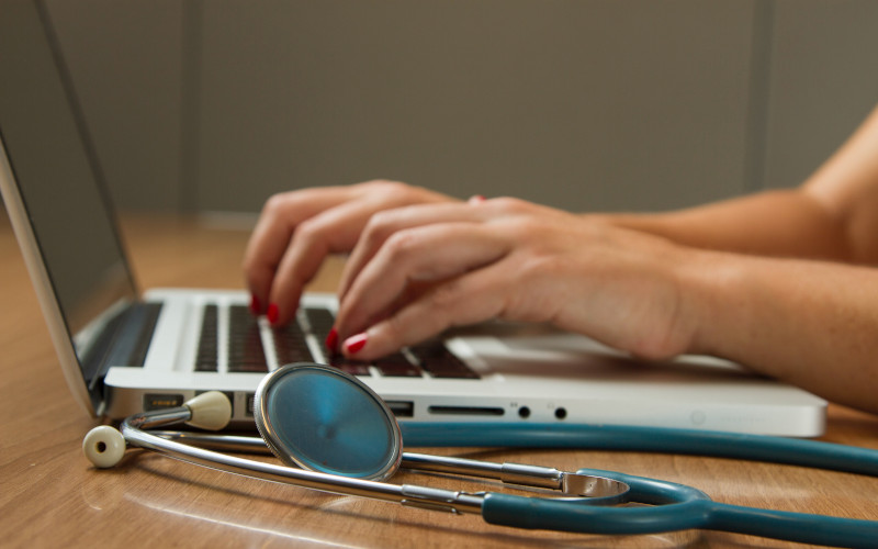 Person typing on a laptop with a stethoscope on the desk